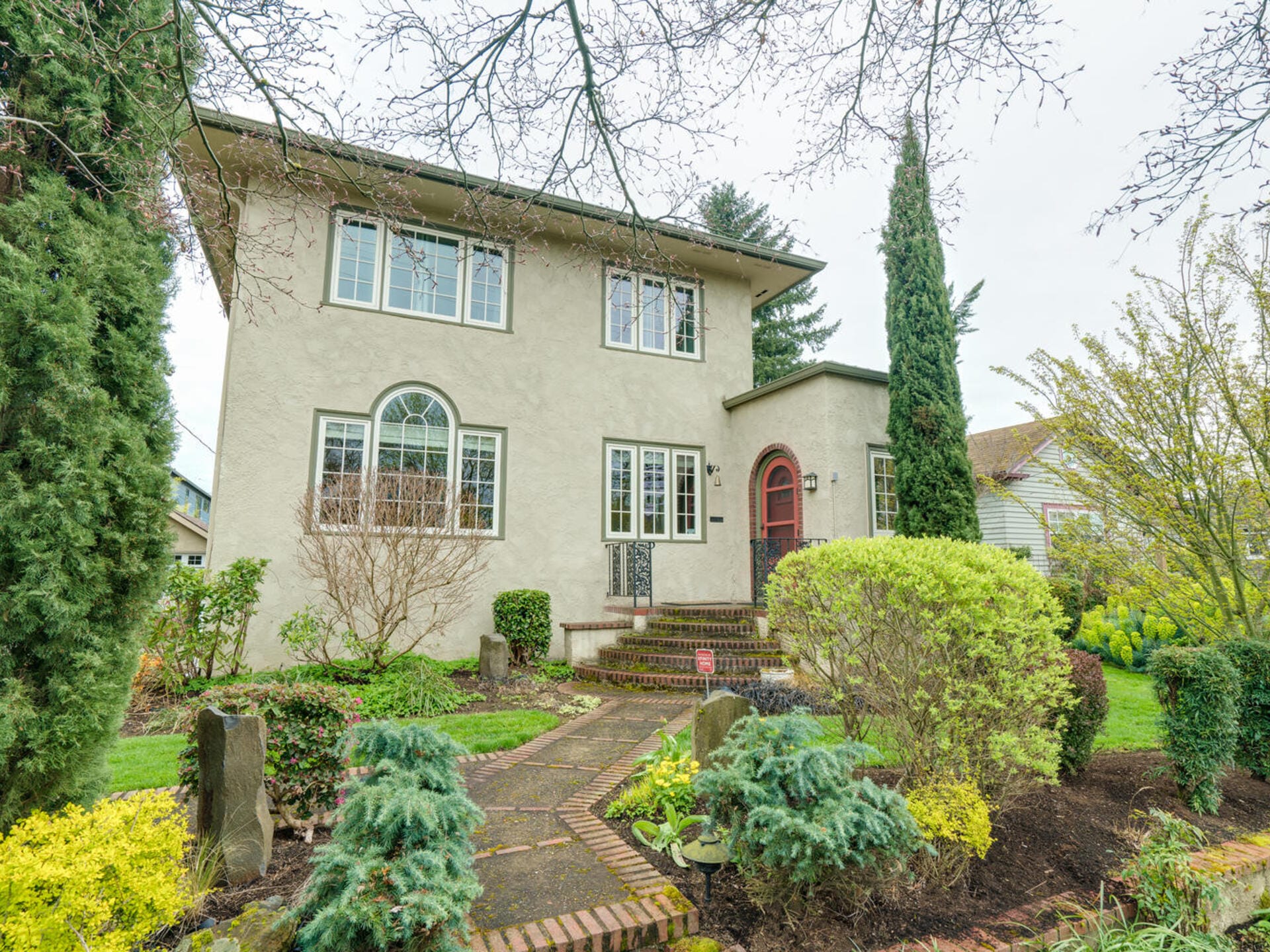A two-story house with a light-colored exterior, surrounded by lush greenery and tall trees. A brick pathway leads to the red front door, with shrubs and well-maintained landscaping in the garden. The sky is overcast.