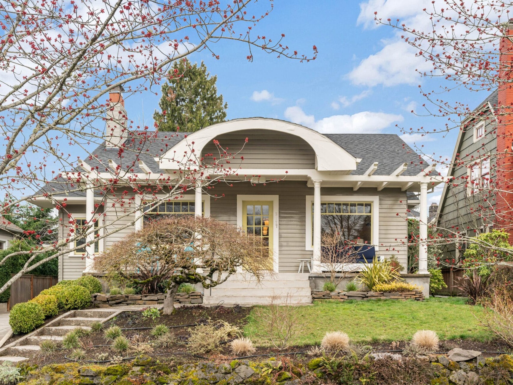 A charming beige house with a porch and white columns, surrounded by a landscaped yard with pruned bushes and bare-branched trees. The sky is blue with a few clouds. A stone pathway leads to the entrance.