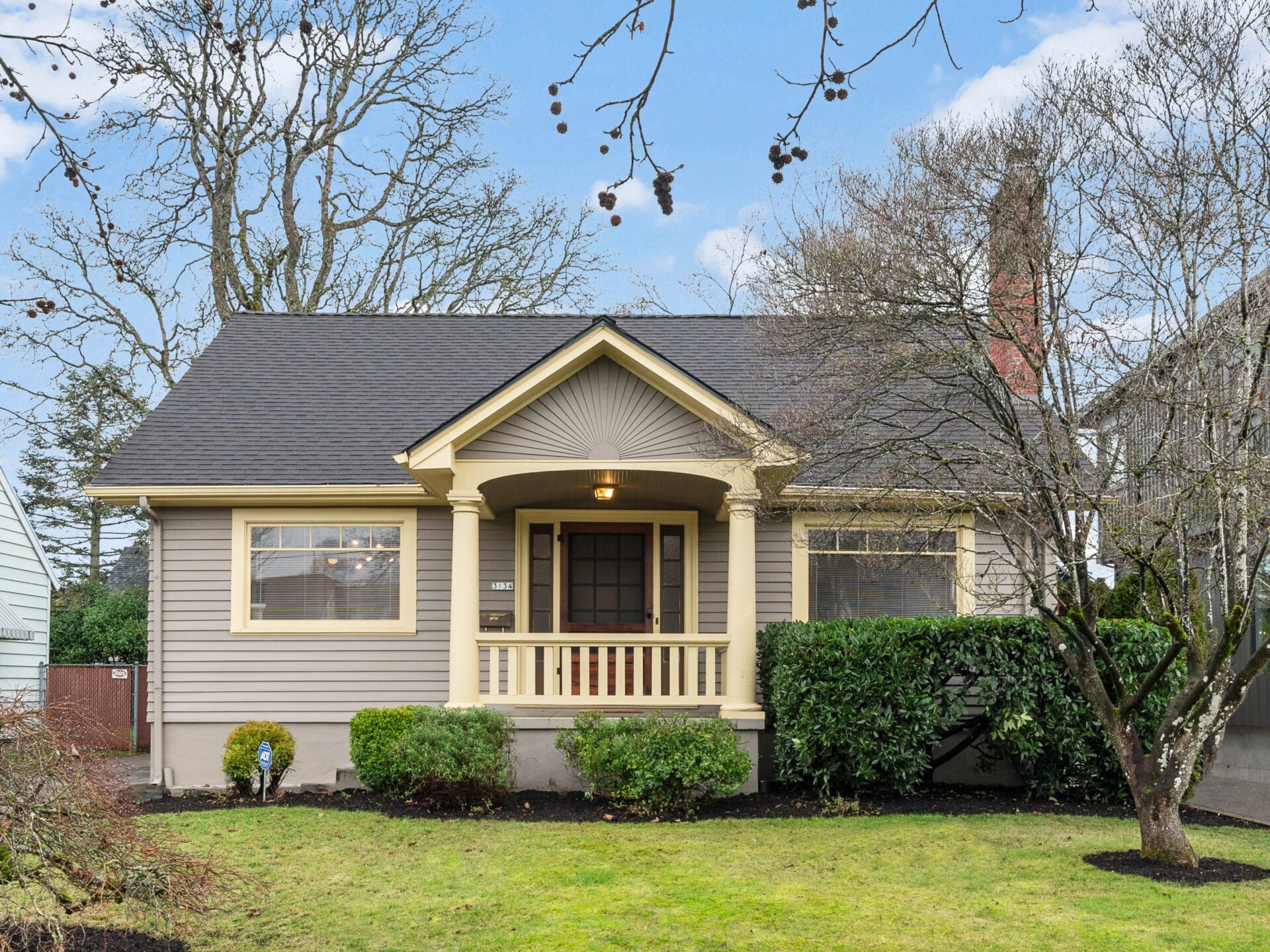 A cozy, single-story house with light gray siding and dark gray roof. The front features a small porch with decorative columns, surrounded by neatly trimmed shrubs and a lawn. Leafless branches frame the scene under a partly cloudy sky.