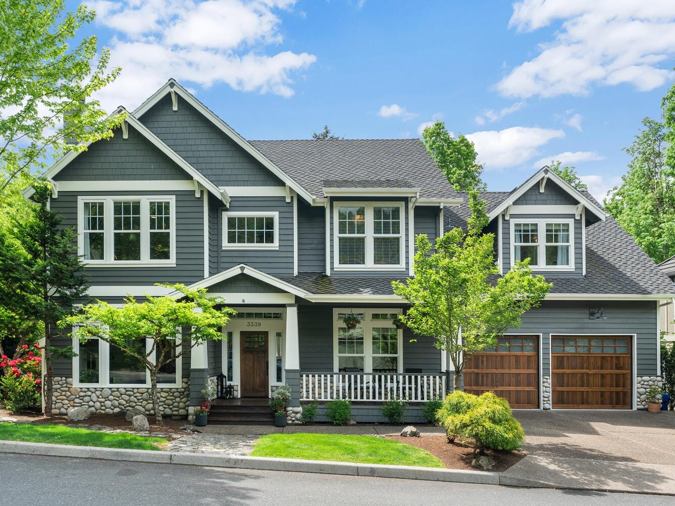 A two-story house with dark gray siding, white trim, and a stone facade. It features large windows, a front porch with a white railing, and two wooden garage doors. The yard is landscaped with trees and shrubs under a clear blue sky.
