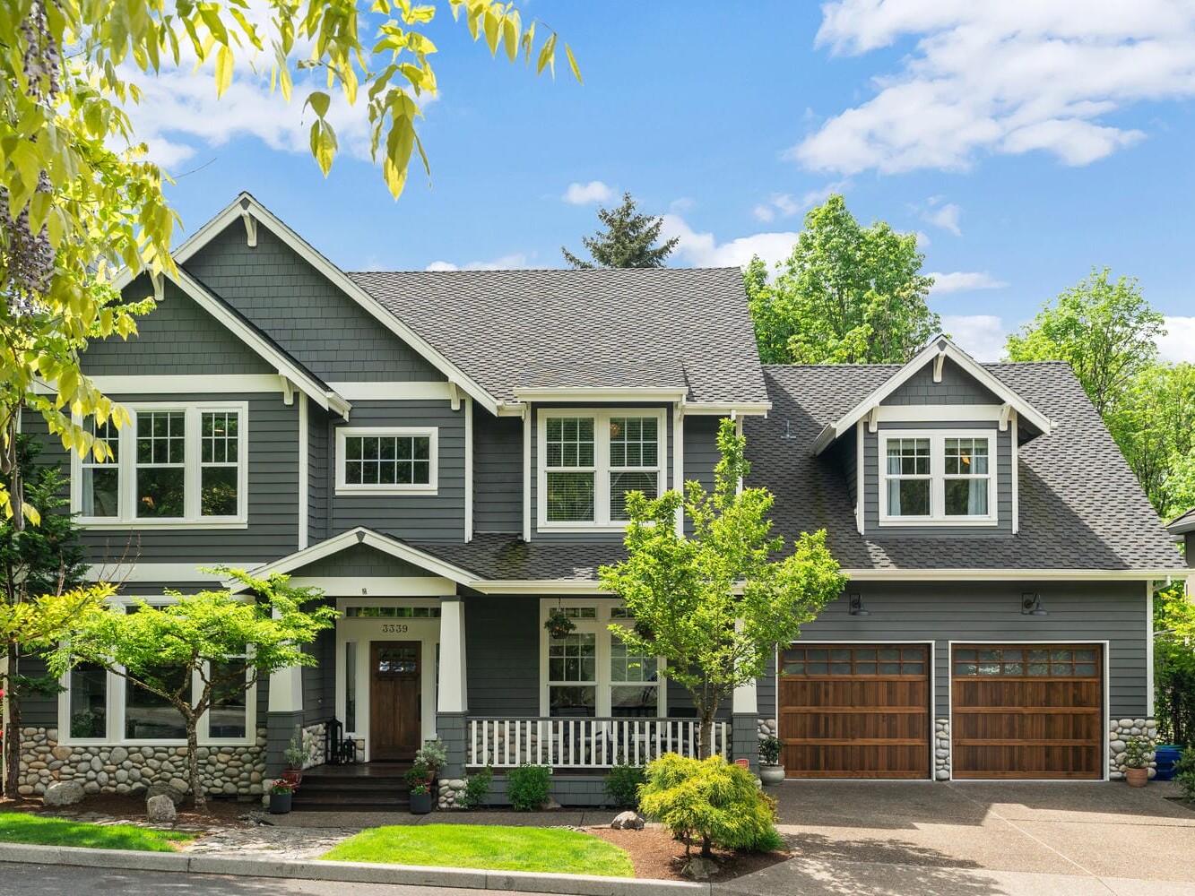 A modern two-story gray house with white trim, a covered porch, and two wooden garage doors. Surrounded by green trees and shrubs, the house has large windows and a well-maintained front yard. The sky is partly cloudy.