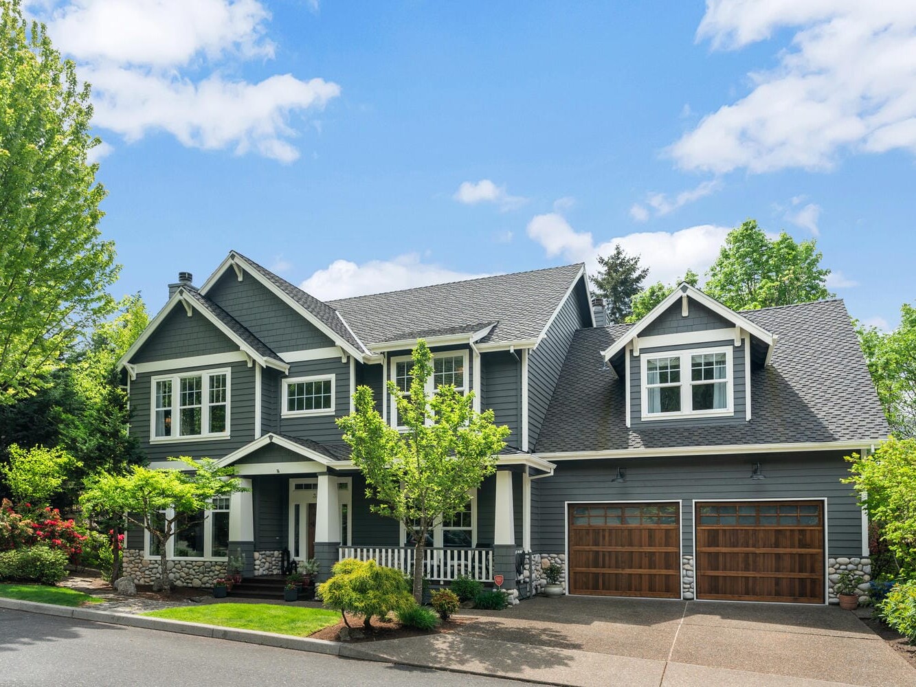 A two-story gray house with white trim features a porch, two wooden garage doors, and a well-maintained lawn with trees and shrubs. The sky above is partly cloudy.