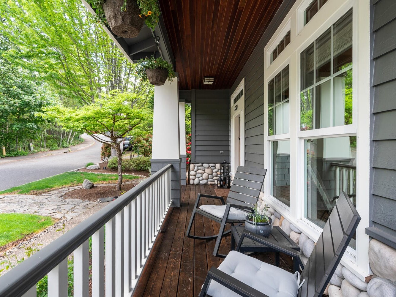 A cozy front porch with dark wooden flooring and two black rocking chairs with cushions. Hanging plants and greenery surround the porch, and a tree-lined street is visible in the background.