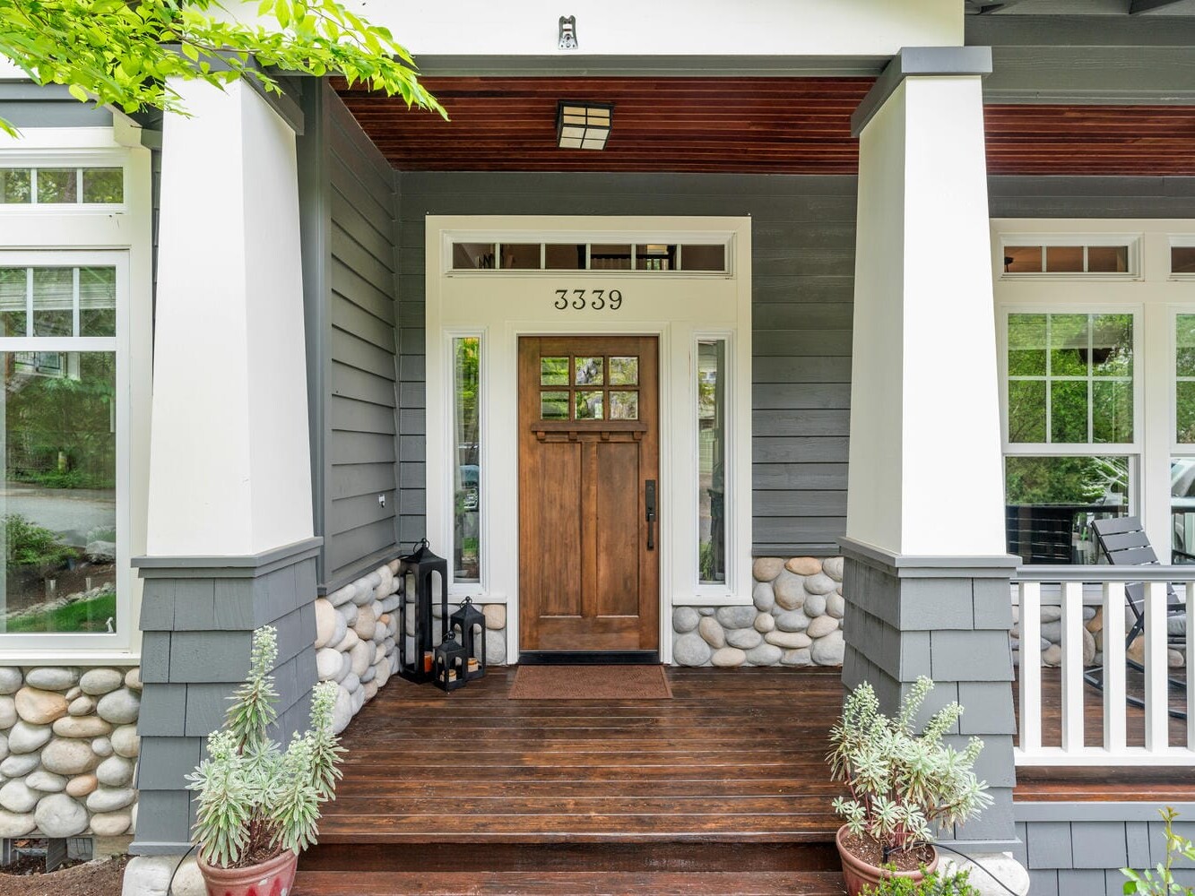 Front porch of a house with a wooden door, flanked by white-trimmed windows. The exterior features grey siding and decorative stonework. Two potted plants sit on the wooden floor, and a pair of lanterns is placed near the door. House number 3339 is displayed above.