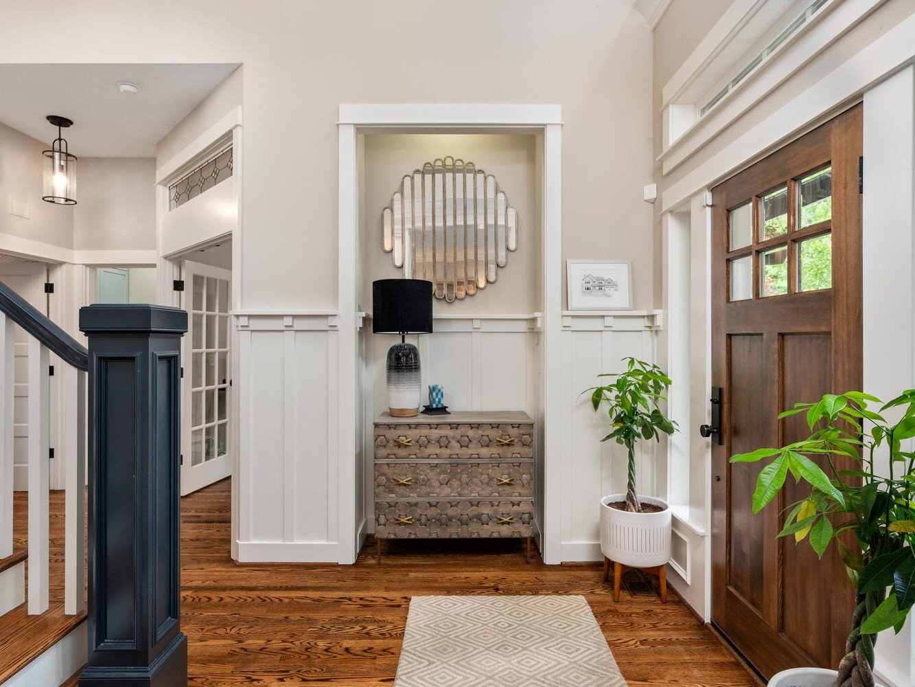A stylish entryway with a wooden floor and a wooden front door. Theres a patterned rug, a potted plant, a chest with a lamp, and an ornate mirror on the wall. White wainscoting and neutral tones create a welcoming atmosphere.