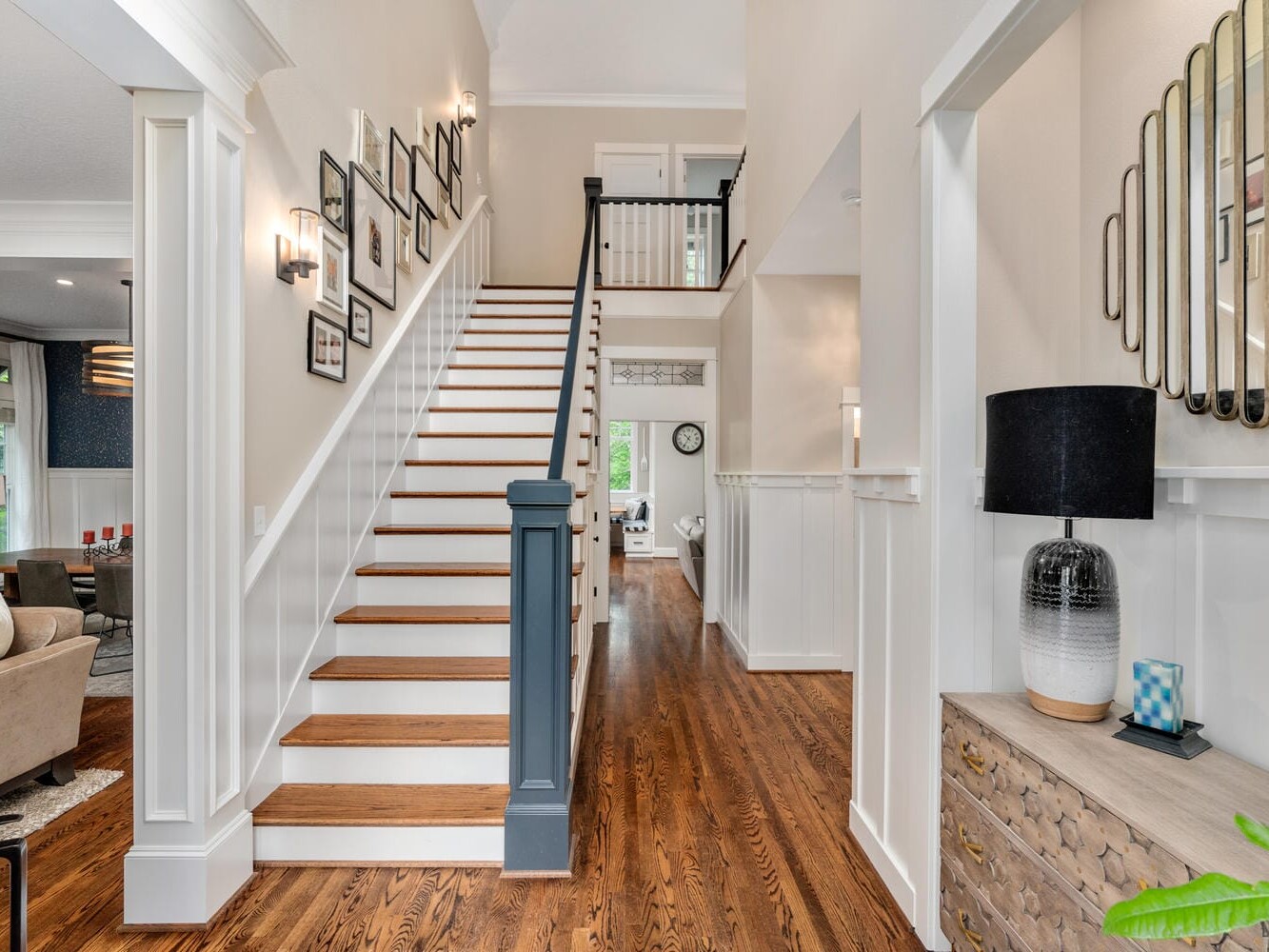 A modern, bright entryway with wooden floors and a staircase leading upstairs. The walls are decorated with framed photos, and a side table with a lamp and mirror is on the right. The living room is visible to the left.