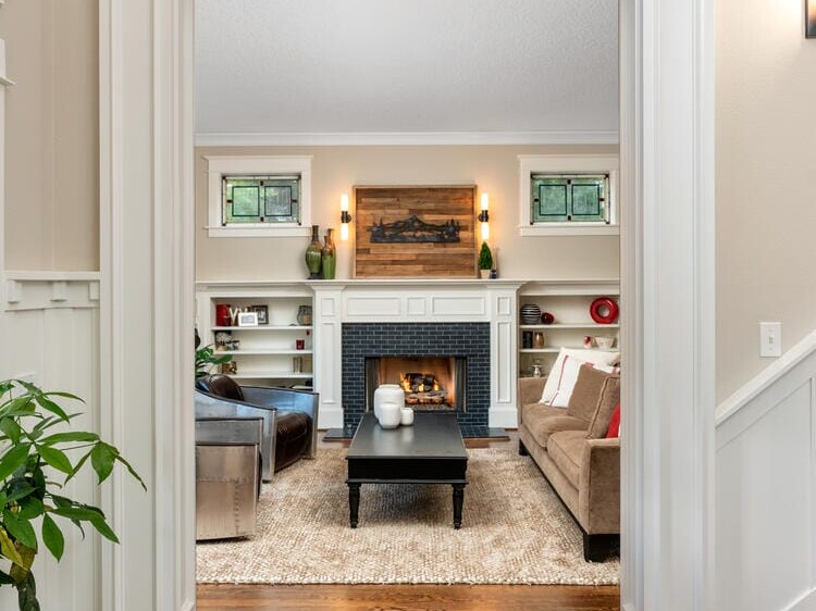 View of a cozy living room through an arched doorway. The room features a fireplace with art above it, surrounded by shelves. A coffee table sits in the center on a textured rug, with comfortable seating around it. Elegant hardwood floors and plants adorn the space.