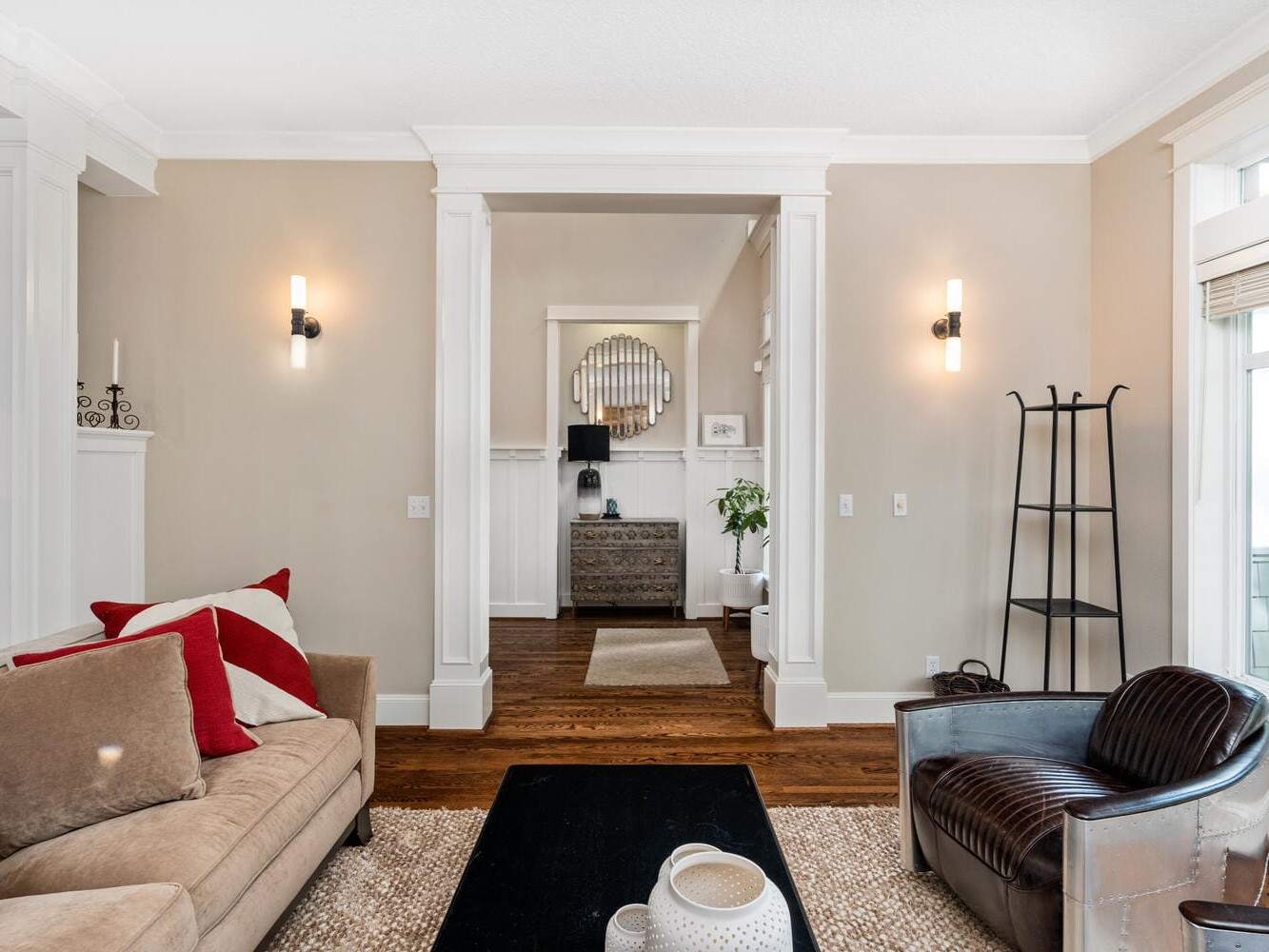 A modern living room with a beige sofa adorned with red and white pillows, a metal armchair, and a black coffee table. The room features light beige walls, wooden flooring, and a view towards a decorated hallway with a mirror.