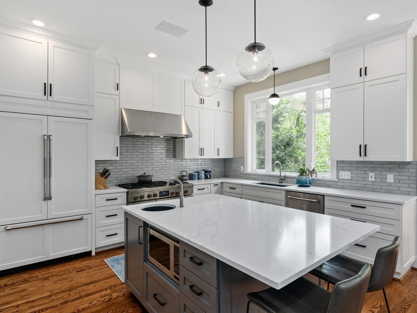 A modern kitchen with white cabinets, a stainless steel stove and range hood, light gray subway tile backsplash, and a large island with a marble countertop. The room has hardwood floors and pendant lights hanging above the island.