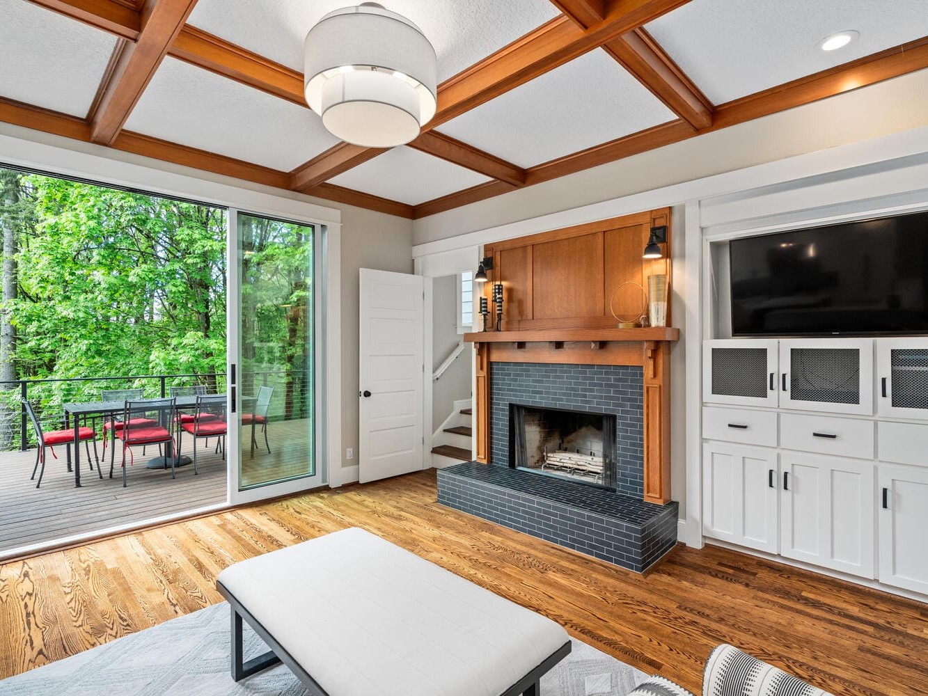 A cozy living room with a large fireplace, wooden ceiling beams, hardwood floors, and built-in cabinets. A sliding glass door opens to a deck with a dining table and chairs, overlooking green trees. Ceiling lighting adds warmth to the space.