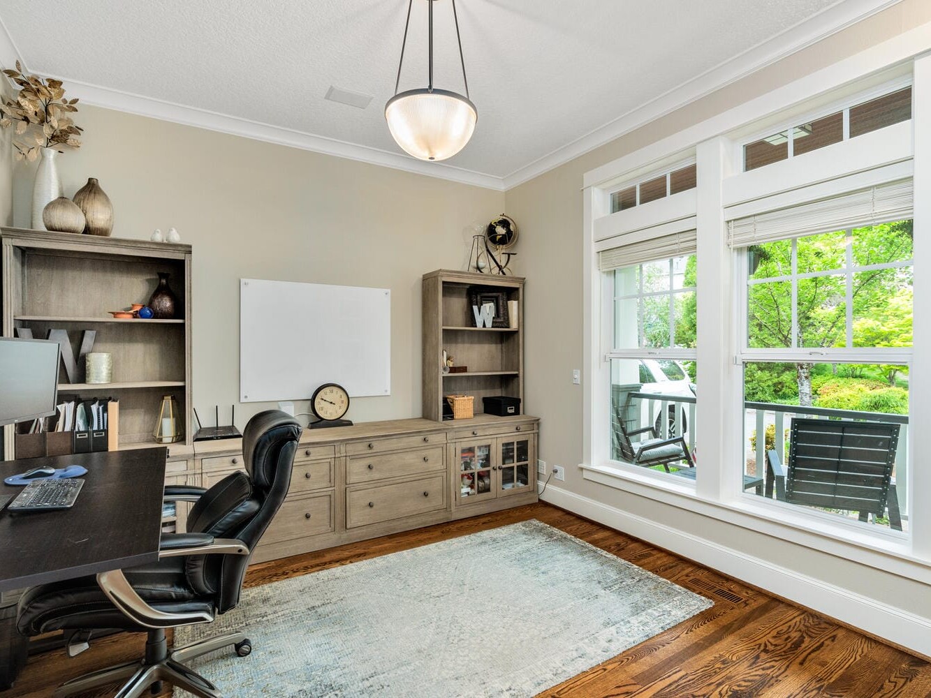 A cozy home office with a large window overlooking greenery. It features a wooden desk with dual monitors, a black chair, shelves with decor and books, a clock, and a neutral-toned rug on a wooden floor. Light beige walls add warmth to the space.