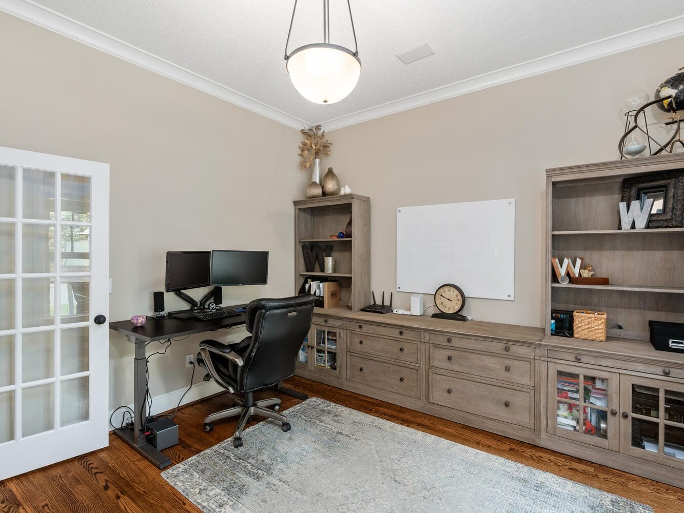 A modern home office with a large desk, ergonomic chair, dual monitors, and a keyboard. Bookshelves and cabinets line the wall. A whiteboard and a decorative globe are visible. The room has a wooden floor and a plush area rug.