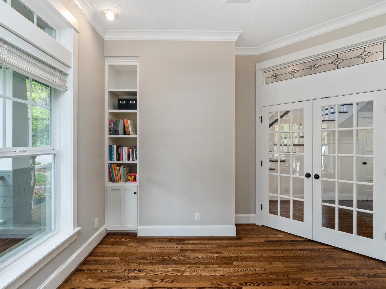 A bright, empty room with large windows and wooden floors. Double French doors with glass panels lead to another area. A built-in white shelf on the left wall holds books and storage boxes. Natural light fills the space.