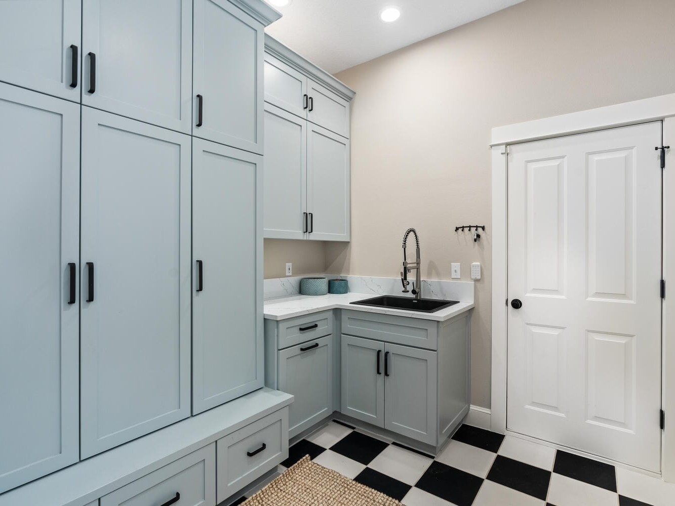 A laundry room with light blue cabinets and drawers, a black sink with a tall faucet, and a countertop. A white door and beige walls complete the room. The floor features a black and white checkered pattern.