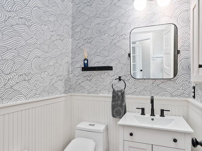 A small bathroom with wave-patterned wallpaper, a white vanity with a black faucet, and a rectangular mirror under two globe lights. A modern white toilet is set against beadboard paneling. A black shelf holds toiletries above the sink.