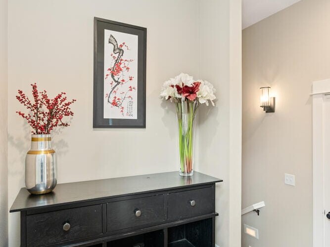 A small hallway features a black console table with open storage, adorned with a vase of red berries and a tall arrangement of white flowers. Above, a framed artwork decorates the beige wall. Wooden floors add warmth to the space.