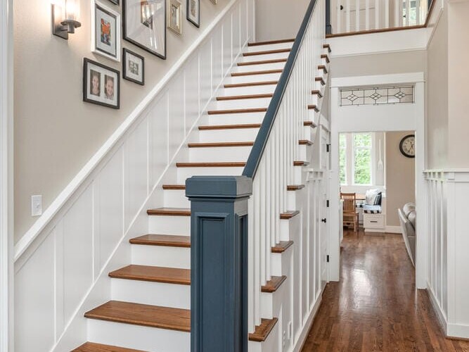 A wooden staircase with white railings and black handrail leads to an upper level. Framed pictures adorn the wall. The space is well-lit with wall sconces, and the polished wooden floor extends to adjacent rooms.