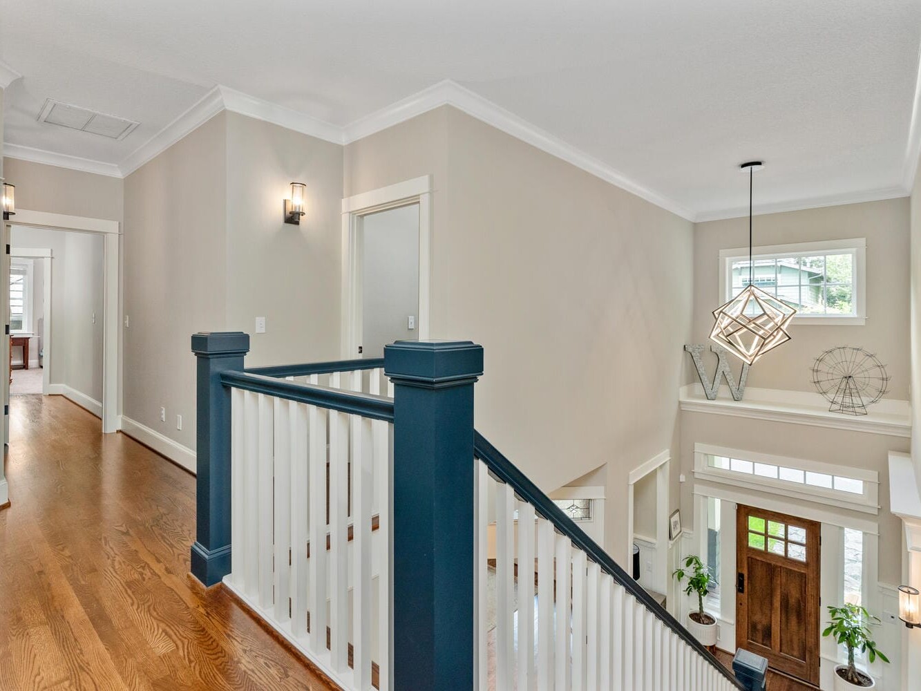Spacious hallway and staircase in a modern home, featuring wooden floors, white walls, and blue accents. The entryway has a large wooden door, plants, geometric decorations, and overhead lighting fixtures. Natural light filters through the windows.