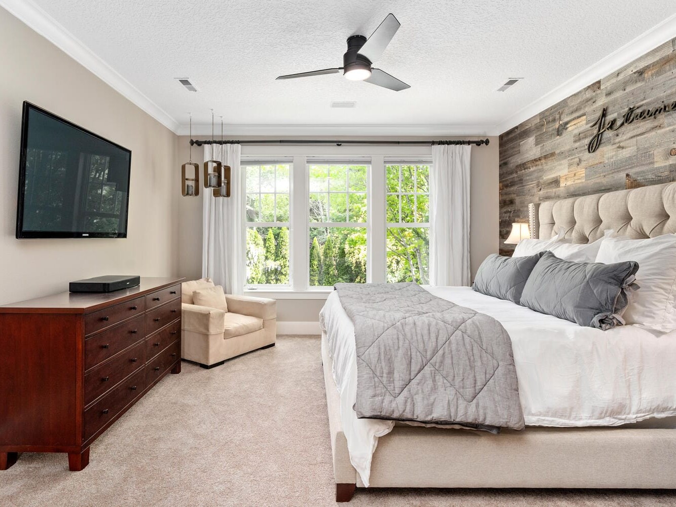 A bright bedroom features a large bed with gray bedding, a tufted headboard, and a rustic wooden accent wall. A ceiling fan hangs above, and a flat-screen TV is mounted on the opposite wall. Large windows let in natural light, illuminating the room.