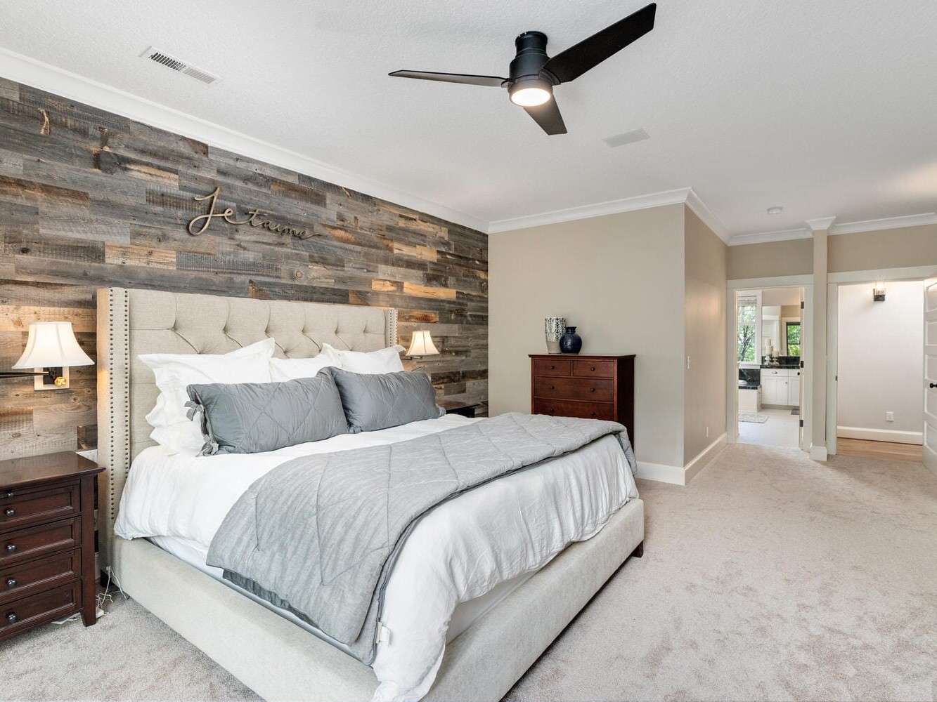 A cozy bedroom with a plush bed featuring gray and white bedding against a rustic wood accent wall. Two wooden nightstands with lamps flank the bed. A ceiling fan hangs above, with a hallway visible leading to a well-lit bathroom.