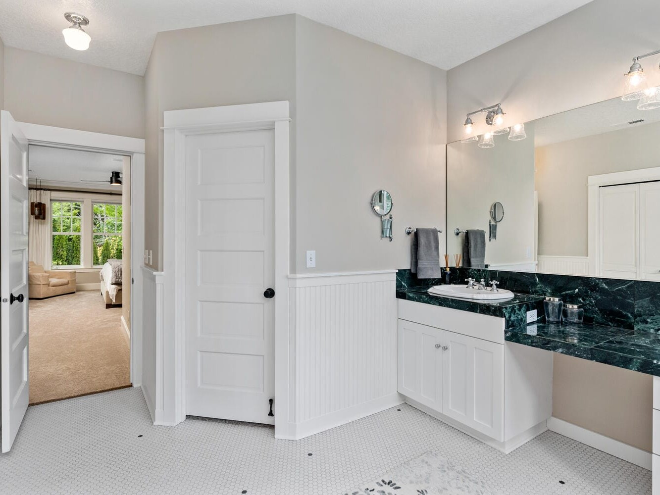 A bright bathroom with white paneled walls and a black marble countertop. It features a large mirror, a sink with silver fixtures, and wall-mounted lights. A door leads to a carpeted room with large windows and a cozy chair.
