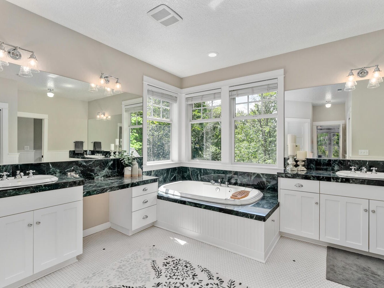 A bright bathroom with large windows, featuring white cabinetry, dual sinks, and a black marble countertop. A built-in corner bathtub with a view of lush greenery outside. The floor is white with a decorative floral pattern.