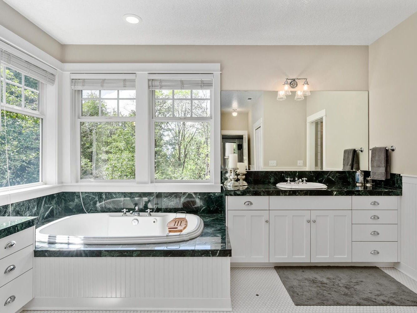 Bright bathroom with a large window, green marble around the bathtub, white cabinetry, and a mirror above the sink. A gray mat is on the tiled floor, and soft natural light fills the room.