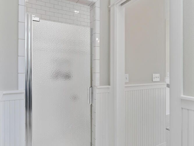 A bathroom with a white tiled shower featuring a glass door. The walls are light grey above white wainscoting. The floor has small hexagonal white tiles. The room appears bright and clean with ample natural light.
