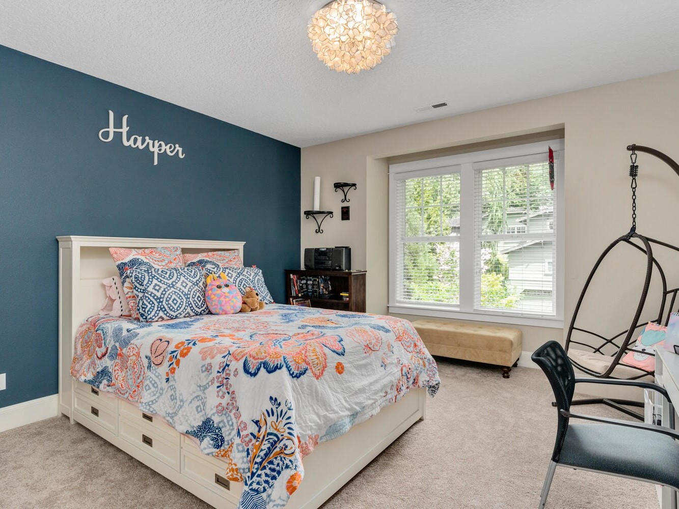 A cozy bedroom features a bed with a colorful quilt, a dark blue accent wall, and a small seating area under a large window. A light fixture hangs from the ceiling, and a chair and desk are on the right side.