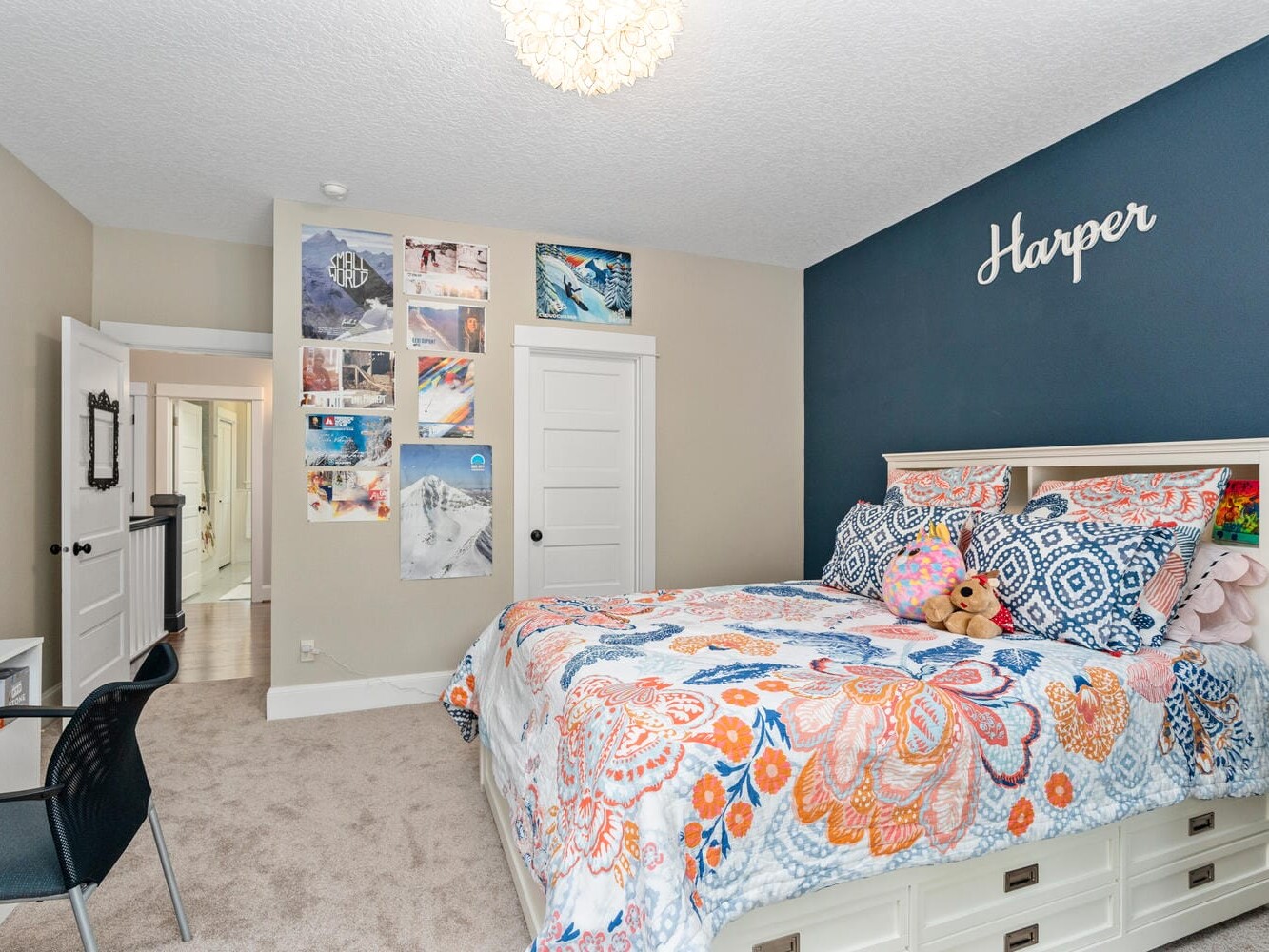 A bedroom with a bed featuring a colorful floral quilt and several pillows, including a teddy bear. A black wall at the head of the bed displays the name Harper. The room includes wall art, a chair, and an open door leading to a hallway.