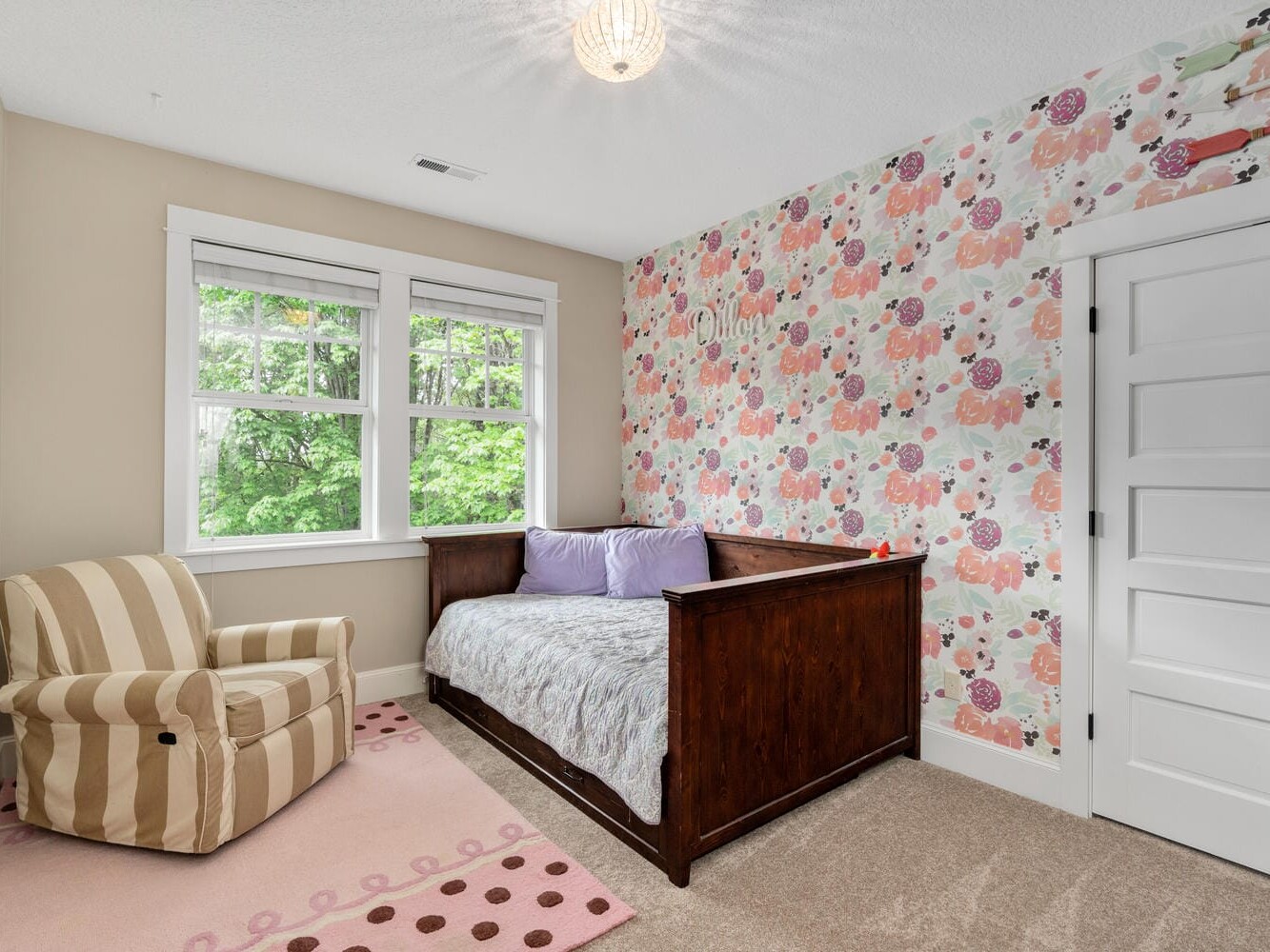 A cozy childrens room featuring a floral accent wall, a dark wooden daybed with purple pillows, a striped armchair, and a pink rug with polka dots. A window provides natural light, and the room has beige walls and a white door.