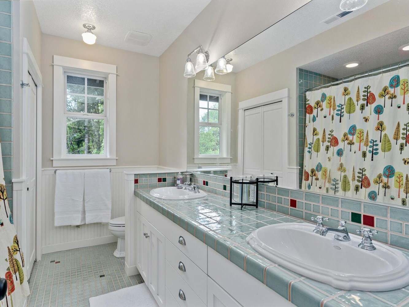 A bright bathroom featuring two sinks with a tiled countertop, a large mirror, and a colorful shower curtain with tree designs. The space is well-lit with natural light from two windows and overhead lighting.