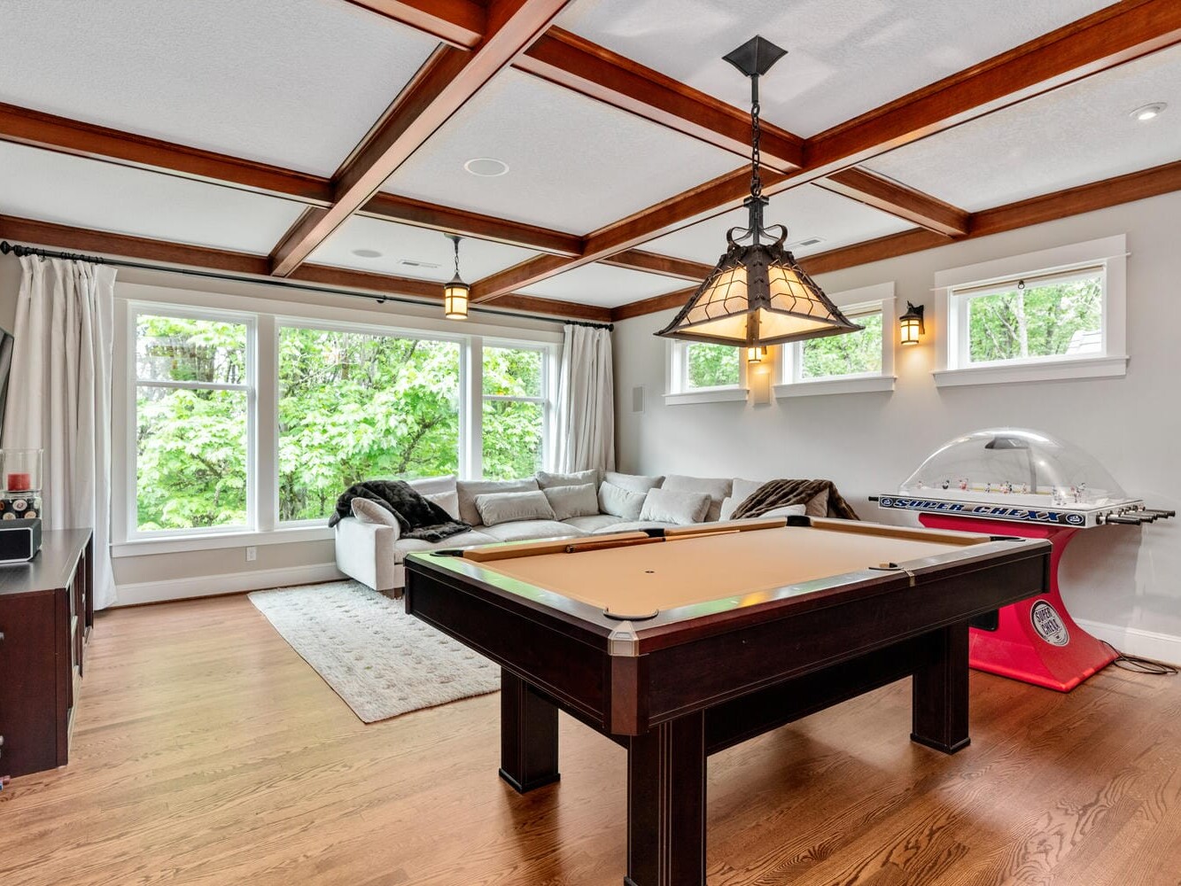 A spacious game room with a pool table centered on hardwood floors. A cozy beige sectional sofa is by large windows with greenery outside. An air hockey table is against the wall, and the ceiling has exposed wooden beams with hanging lights.