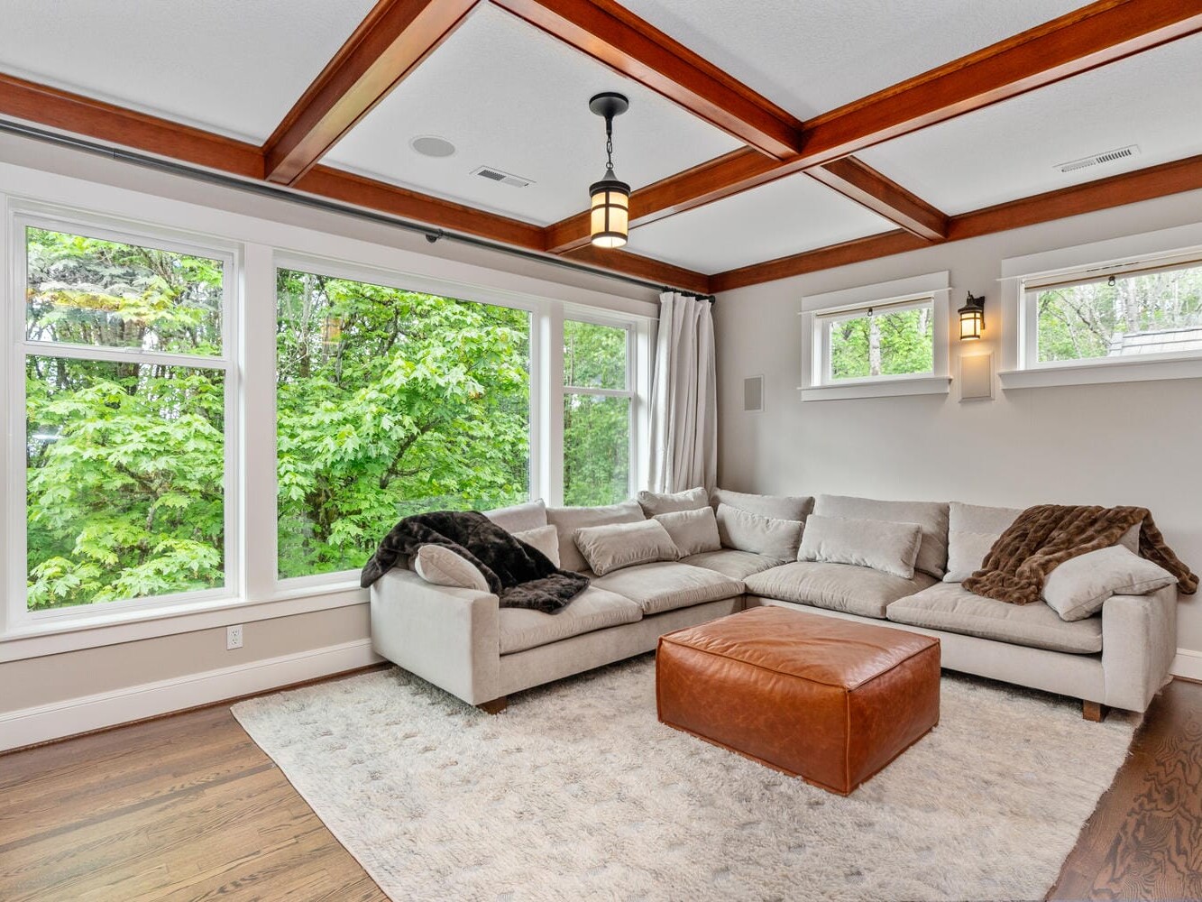 A cozy living room with a large beige sectional sofa and brown throw blankets, facing a window with lush green views. The room features wood beam ceilings, a soft rug, a brown leather ottoman, and two small wall windows with lantern-style lights.