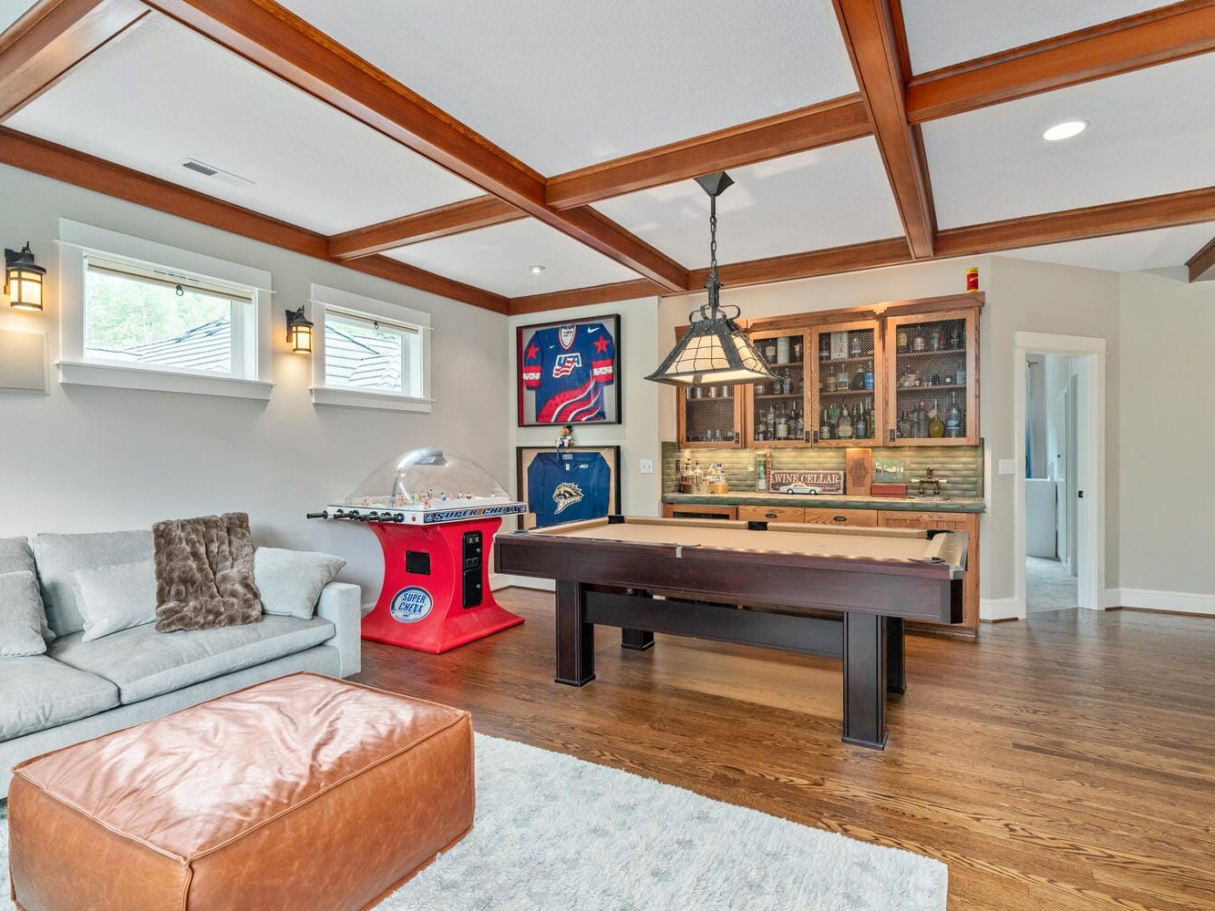 A game room with wooden ceiling beams features a pool table, a foosball table, and a cozy gray sofa. A large ottoman sits nearby on a light blue rug. A wall-mounted cabinet holds various bottles, and sports jerseys are displayed on the wall.