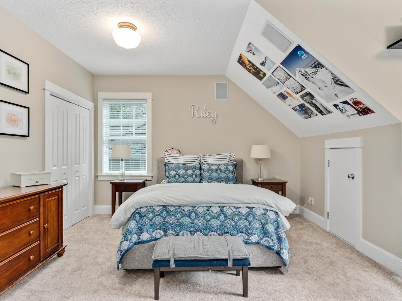 A cozy bedroom with a bed featuring blue patterned bedding and matching pillows. Artwork and photos are displayed on the wall and sloped ceiling. Wooden furniture, including a dresser and nightstand, enhances the warm atmosphere.