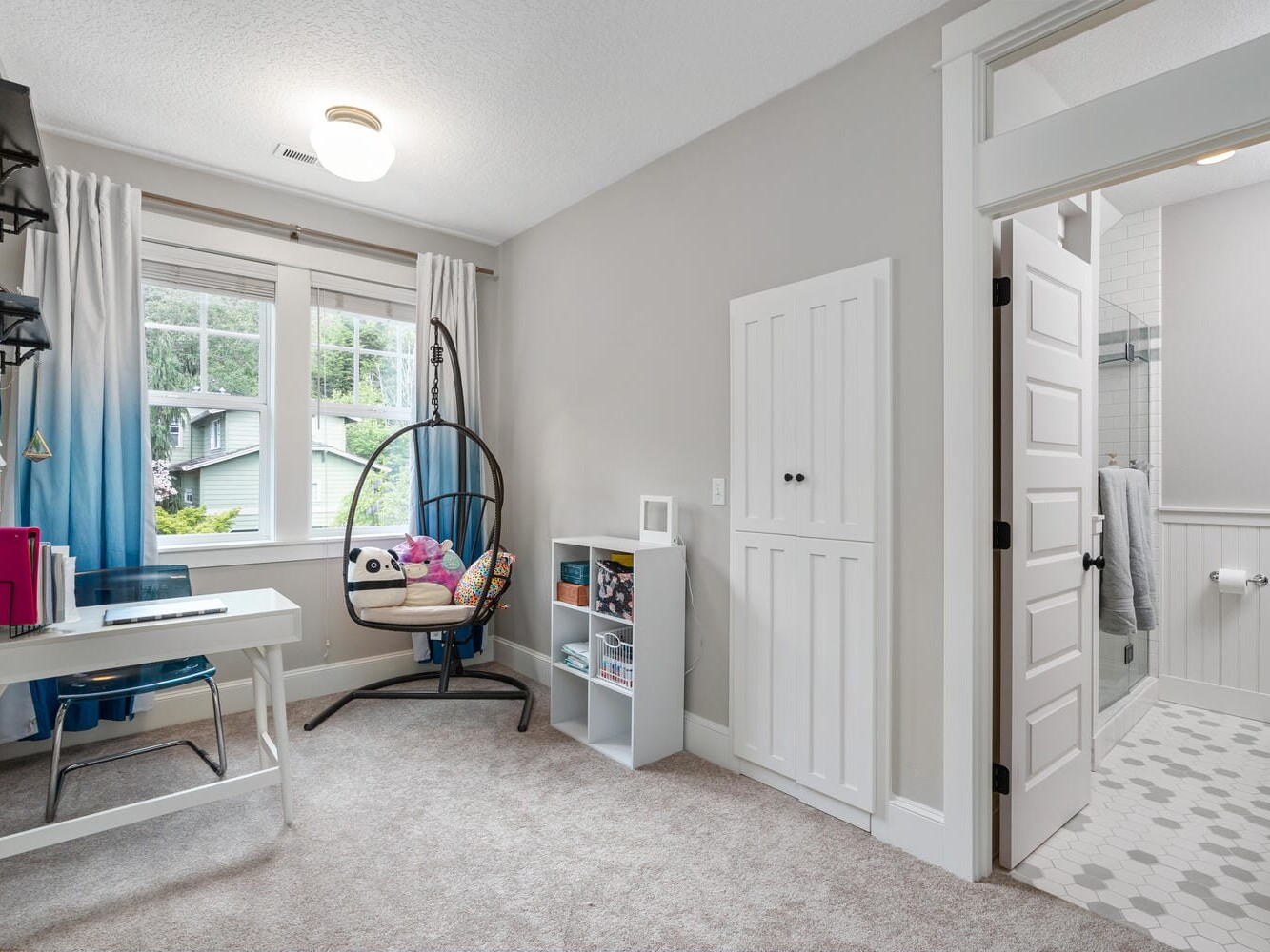 A cozy room with a desk, chair, and hanging chair with a panda cushion by the window. A small bookshelf holds books and toys. A door leads to a bathroom with a hexagonal tile floor. Bright and tidy atmosphere.