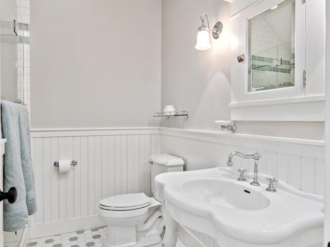 A bright bathroom with a vintage-style sink and mirror. White wainscoting and gray walls complement the hexagonal tile floor. A toilet and towel are visible near a glass shower door, under a small ceiling light.