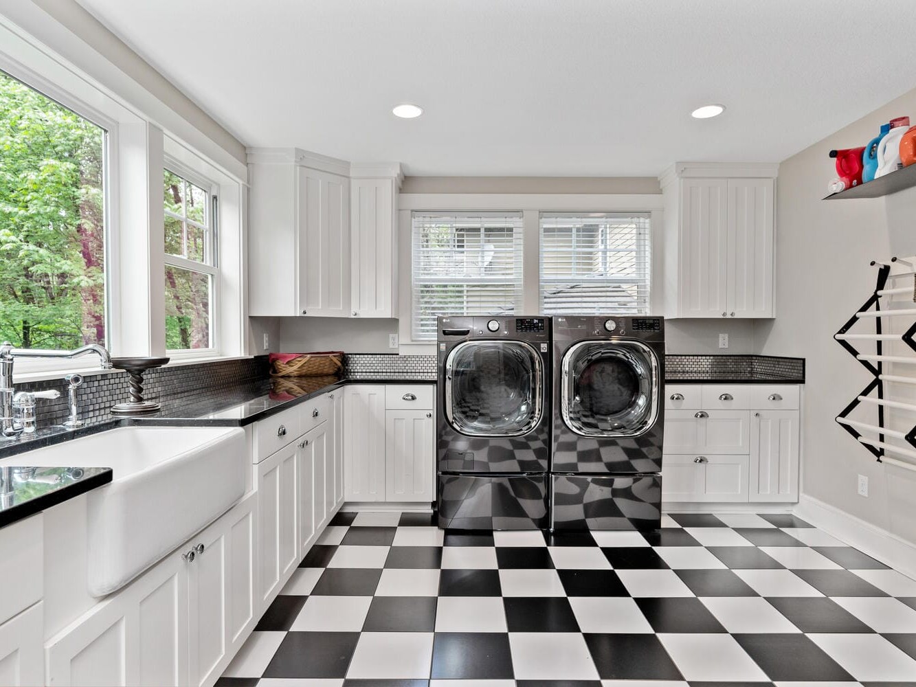 Bright laundry room with white cabinets, black countertops, and black-and-white checkered flooring. Two large washers sit under a window. Shelves with baskets are on the right, and greenery is visible outside through the windows.