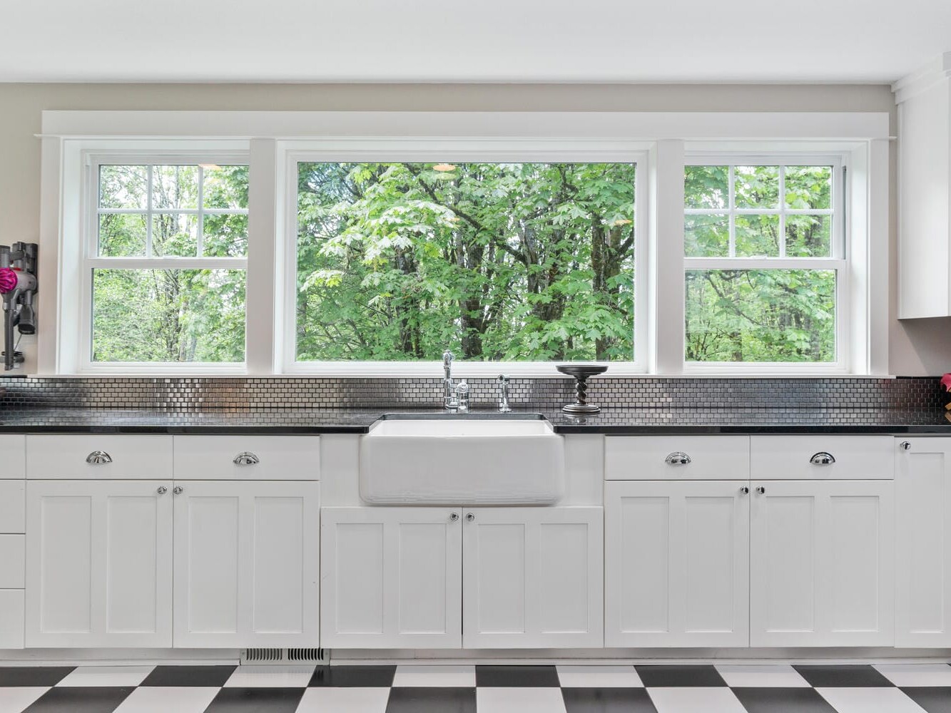 A bright kitchen features white cabinets, a farmhouse sink, and large windows with a view of lush green trees. The room is accented by a black and white checkered floor and silver backsplash, creating a clean and classic look.