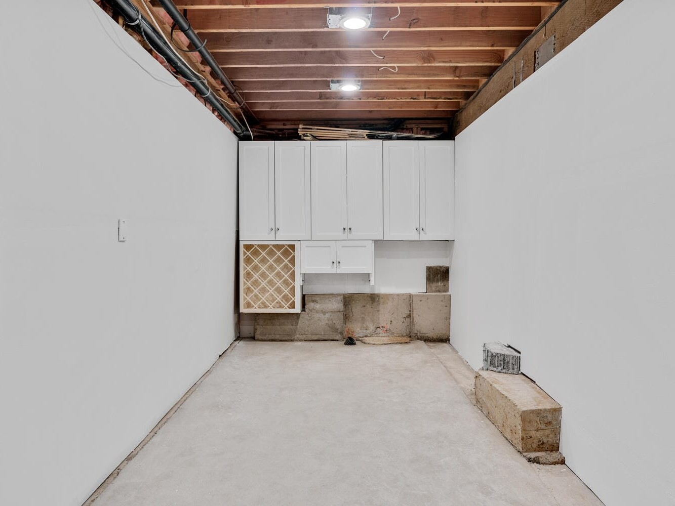 A bare basement room with white walls and an unfinished wooden ceiling. There are white cabinets mounted on the wall, and a small step made of concrete blocks on the right side. The floor is bare concrete.