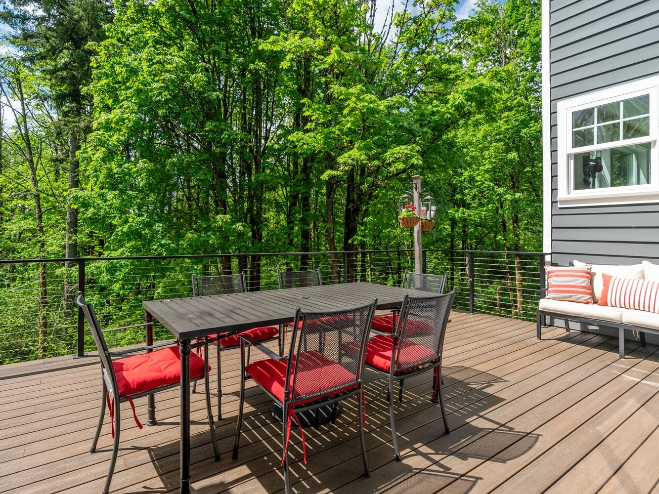 A wooden deck with a dining table and six chairs with red cushions. A gray house wall is visible, featuring a window. A white bench with red and white striped pillows sits nearby. Lush green trees provide a natural backdrop.