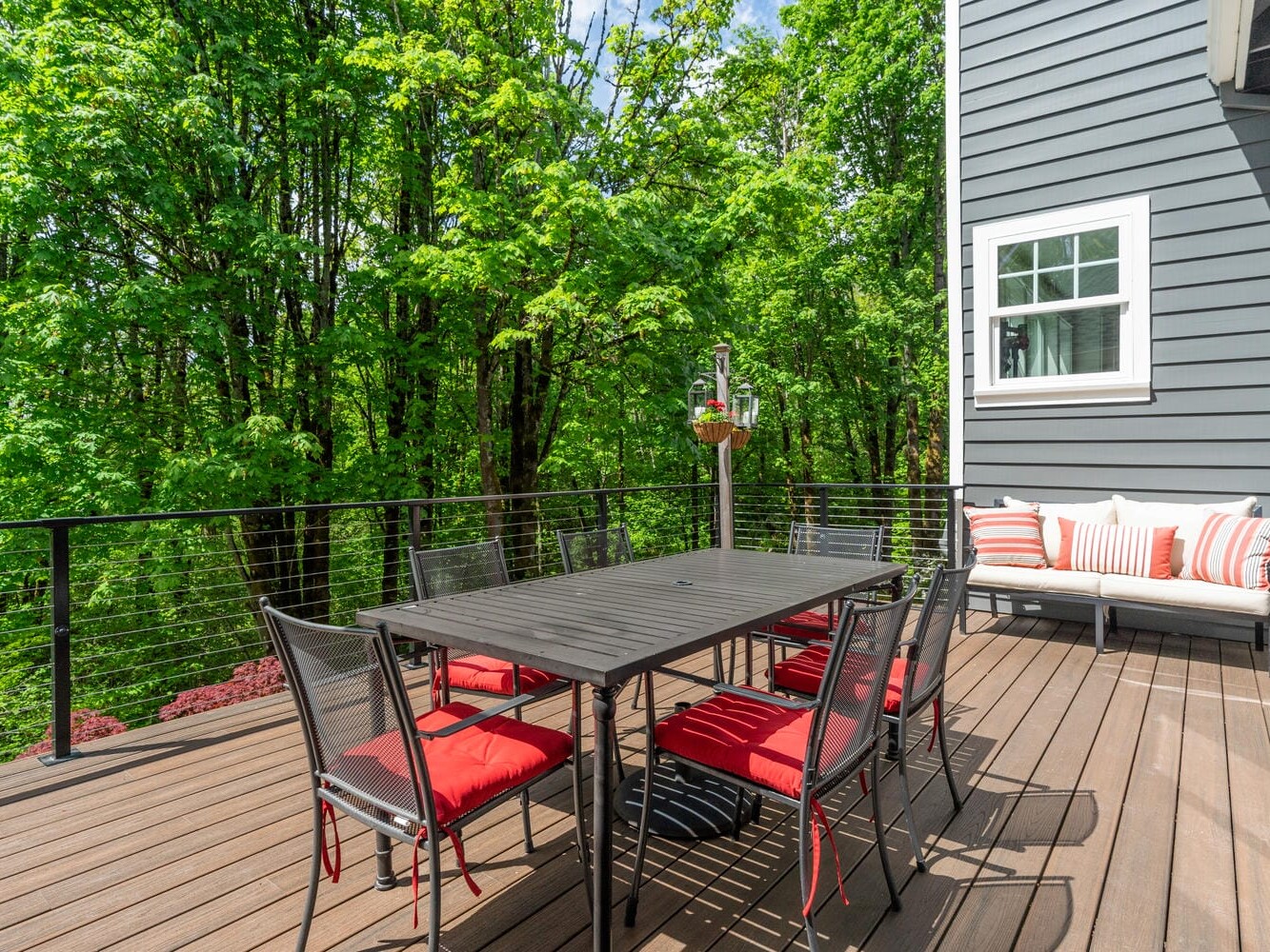 A spacious wooden deck with a black dining table and six chairs featuring red cushions. A nearby cushioned bench with striped pillows is against a gray house wall. Lush green trees surround the area, and the sky is clear.
