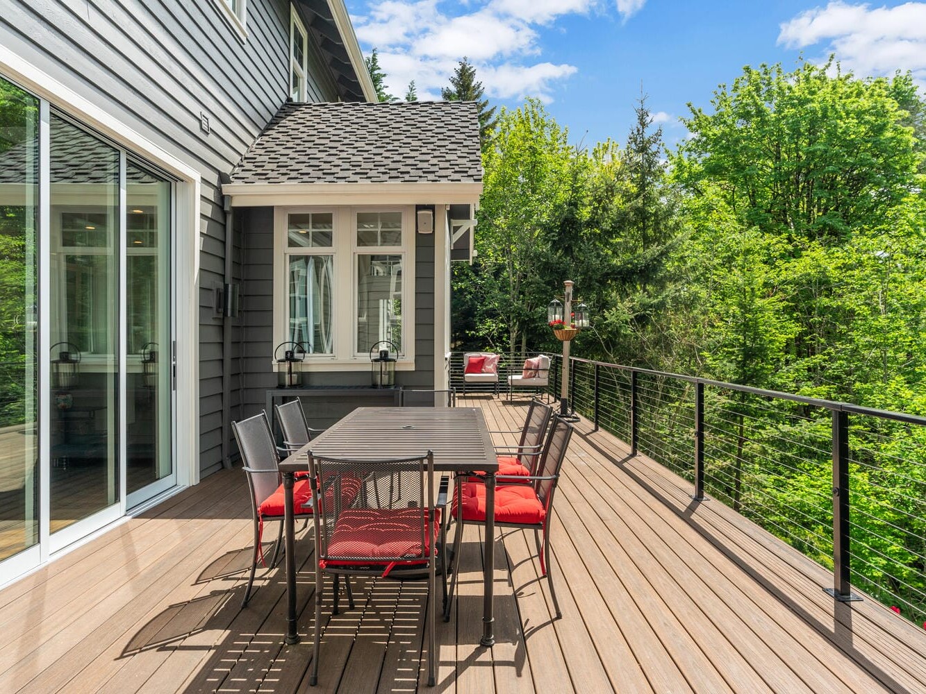 Spacious wooden deck attached to a house, featuring a dining table with red cushioned chairs, a sleek railing, and views of lush green trees under a partly cloudy sky.