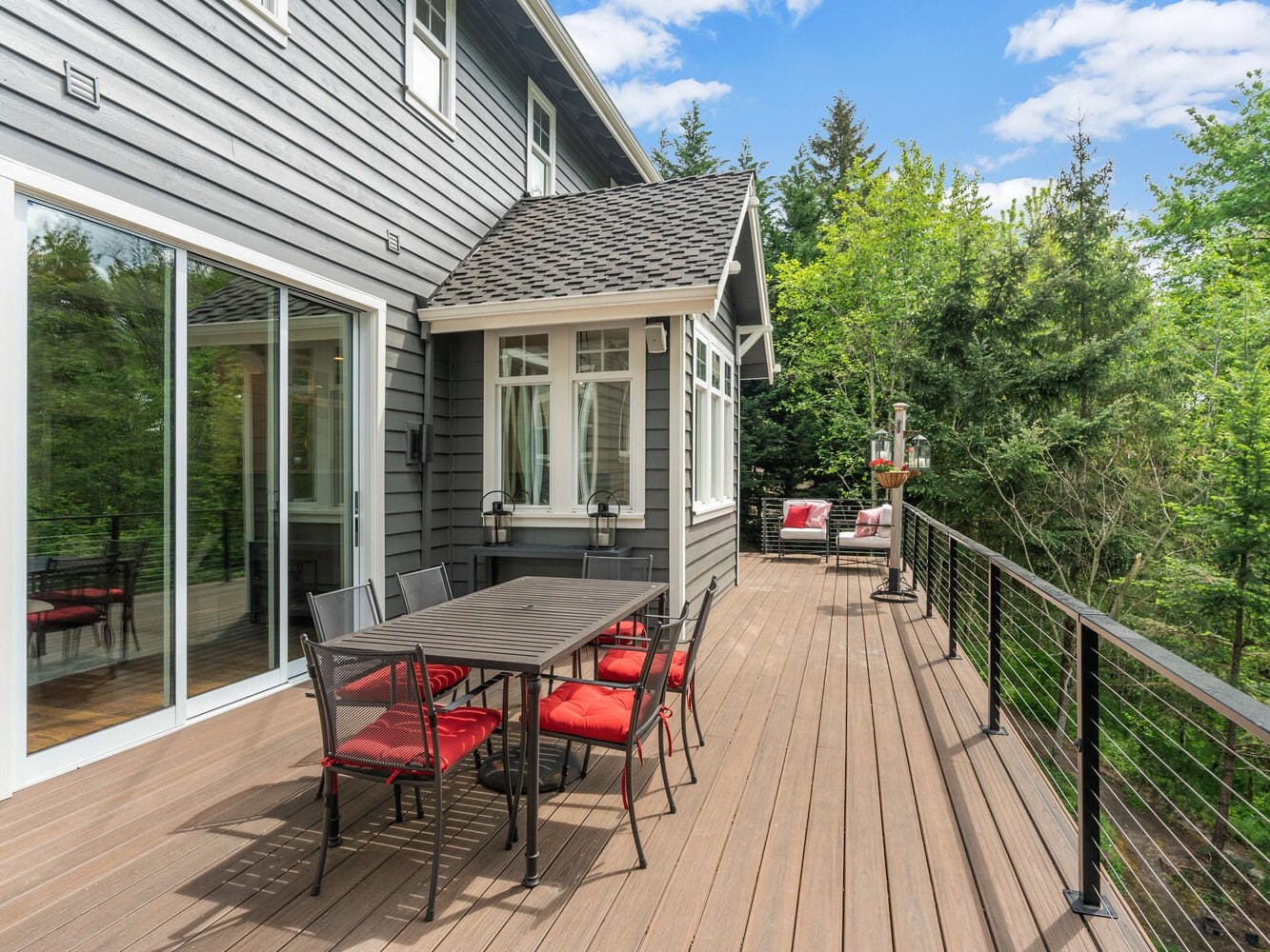 A wooden deck attached to a grey house, featuring a dining set with black chairs and red cushions. There is more seating in the background with red cushions. Lush green trees surround the deck under a blue sky with clouds.