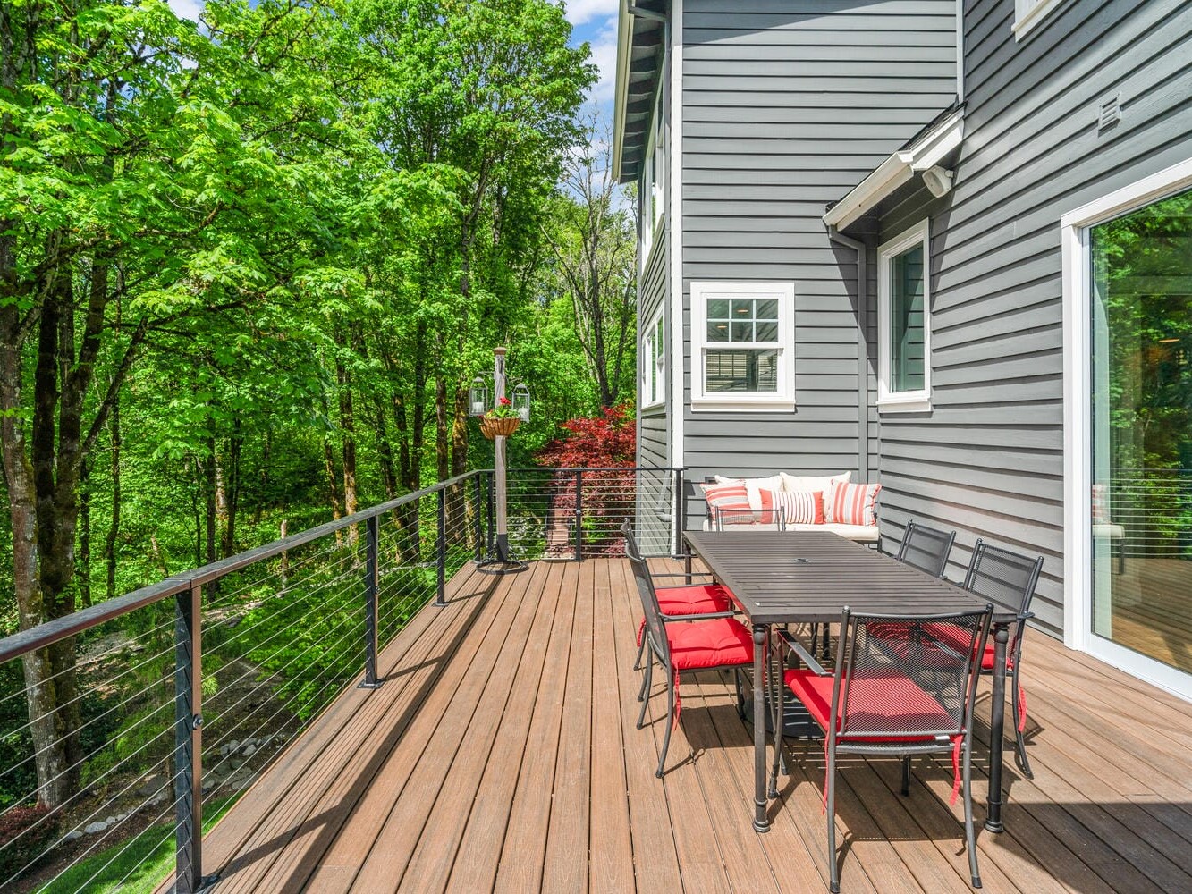 A modern deck attached to a gray house features a rectangular dining table with red cushioned chairs. It overlooks a lush, green forest. The deck has cable railing and a sliding glass door leading inside.
