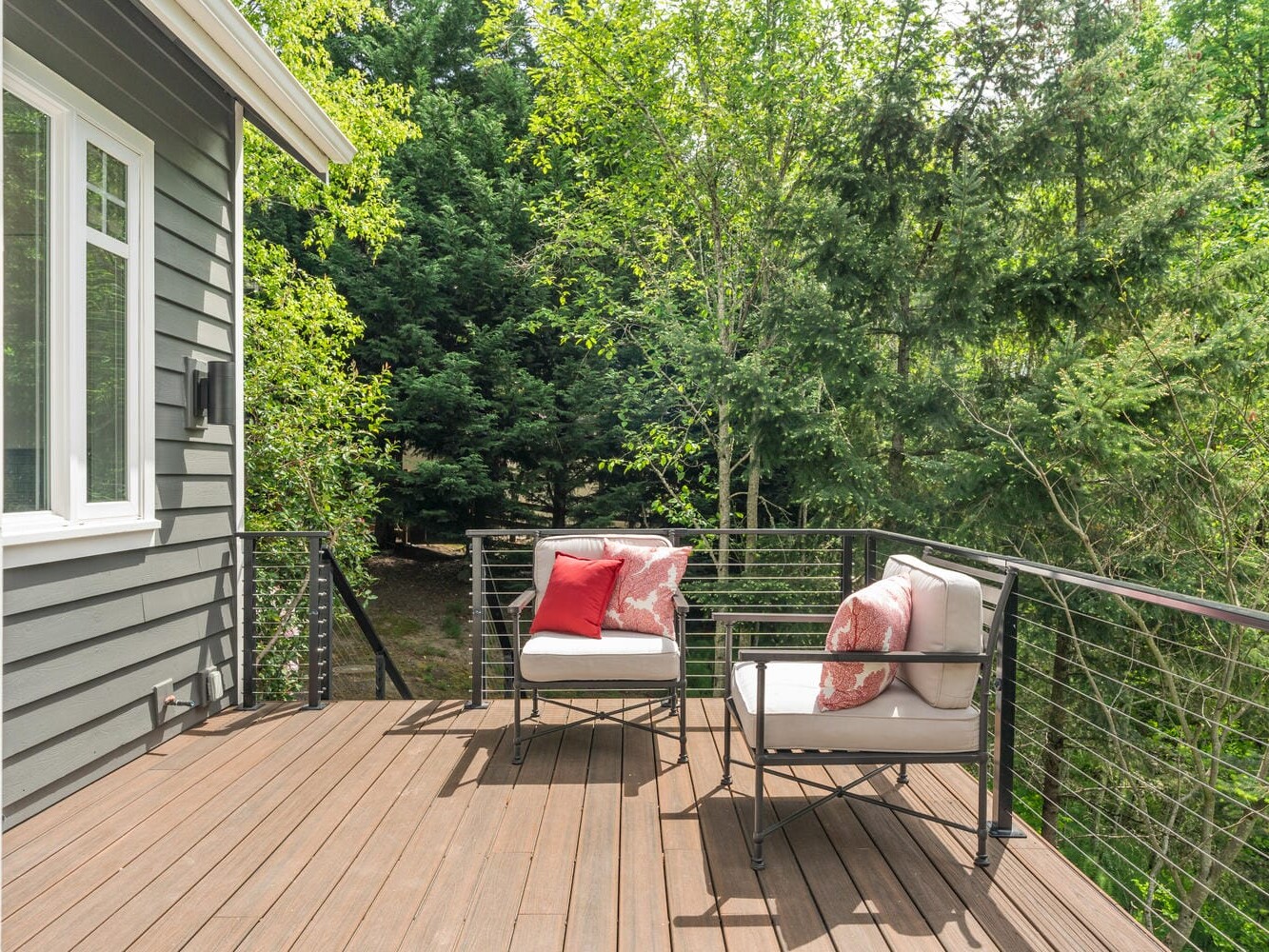 A wooden deck with two cushioned chairs adorned with red and white pillows overlooks a lush, green forest. The deck is attached to a gray house with white trim and surrounded by black metal railings.