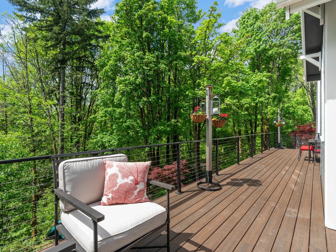 A spacious wooden deck with outdoor seating and a bird feeder overlooks lush green trees under a partly cloudy sky. A cushioned chair with a red pillow is in the foreground, next to hanging flower baskets.