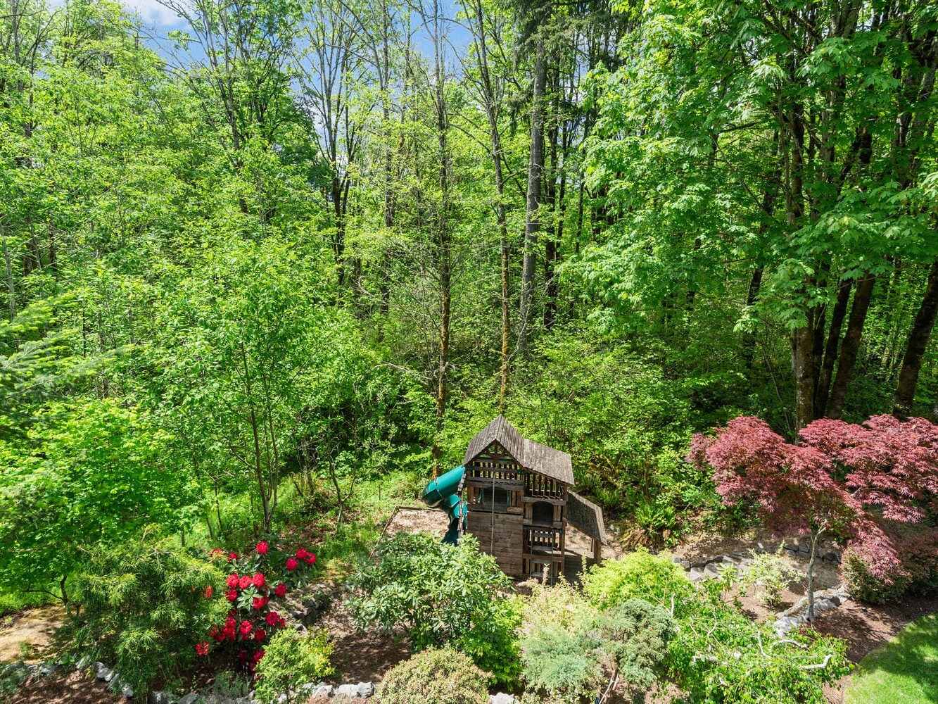 A lush, green backyard filled with trees and bushes, showcasing a wooden playhouse with a slide. Red and pink flowering plants are in bloom, and the sky is clear with patches of blue visible through the canopy.