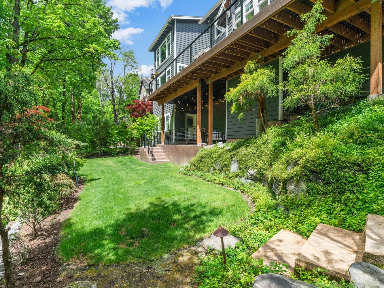 A lush green backyard features neatly manicured grass, a variety of trees and shrubs, and a wooden deck attached to a dark-colored house. Wooden steps lead onto the lawn, creating a serene and inviting outdoor space under a blue sky.
